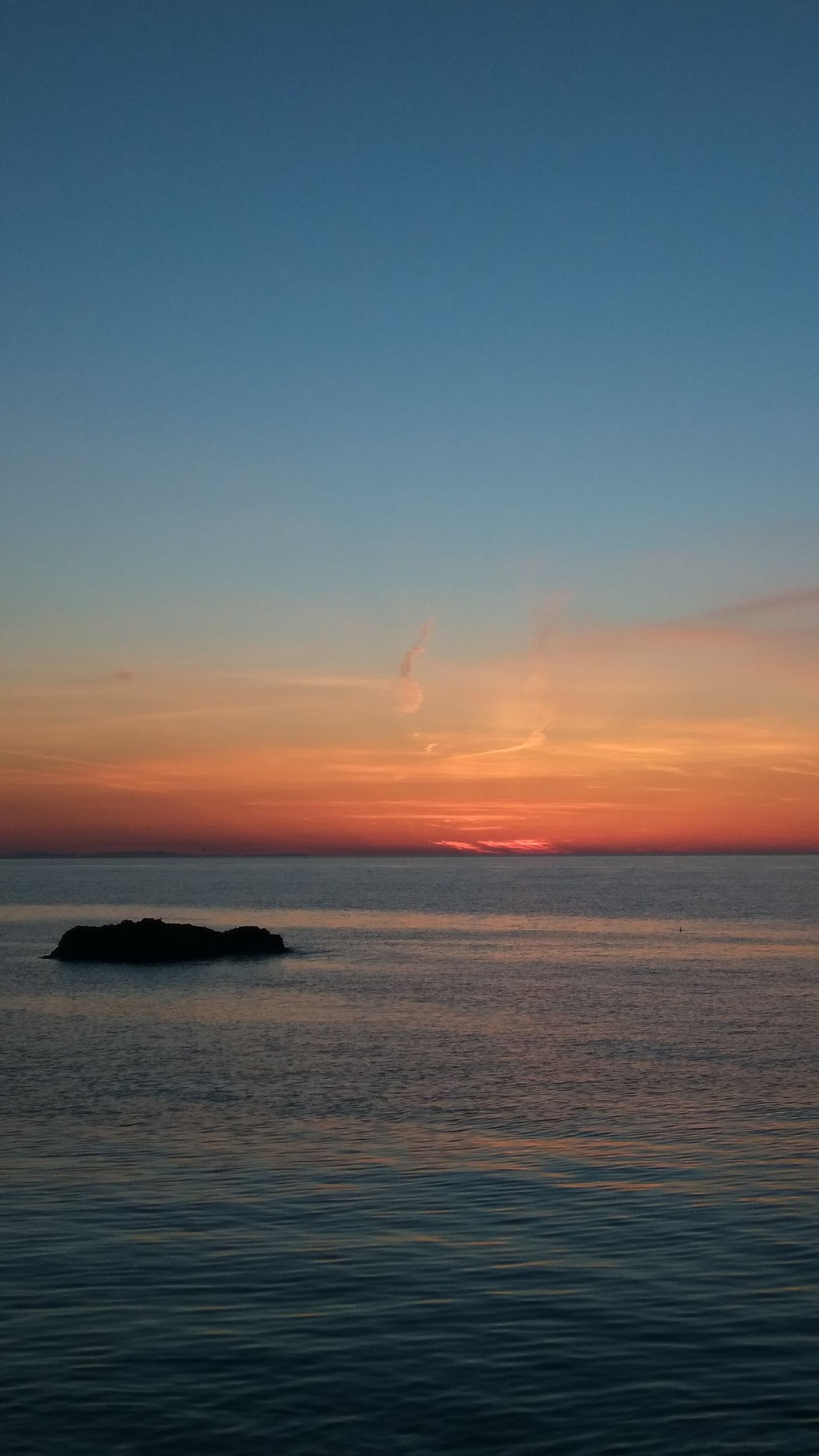 Peaceful ocean sunset with vibrant sky colors and a distant rock in view.